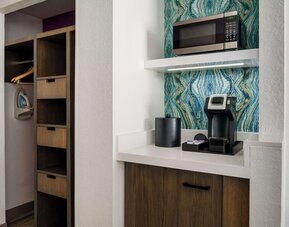 In-room coffee station with a single-serve coffee maker, microwave, ice bucket, storage cabinet, open closet, hangers and ironing board against a decorative blue accent wall.