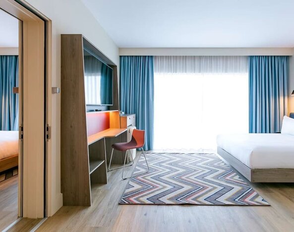 Spacious modern hotel guest room with king-size bed, floor-to-ceiling window with blue curtains, wall-mounted television, work desk and chair, wood flooring, and colorful geometric rug.