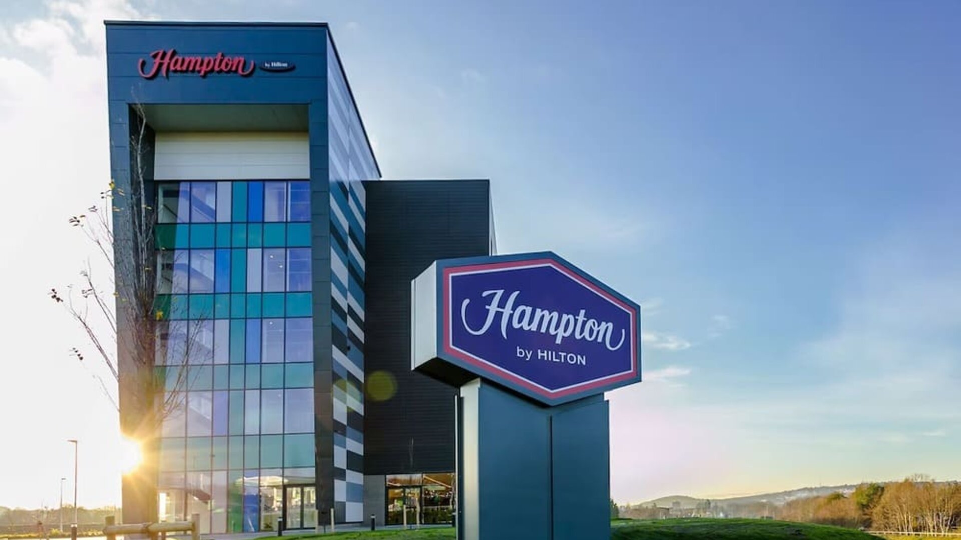Exterior of Hampton by Hilton hotel featuring a modern multi-story glass and dark-panel façade, large freestanding Hampton by Hilton sign, landscaped grounds, and blue sky.