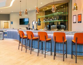 Modern Hampton by Hilton hotel bar with a long stone countertop, bright orange high-back bar stools, geometric pendant lights, beer taps, bottle displays, and illuminated shelving.