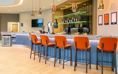 Modern Hampton by Hilton hotel bar with a long stone countertop, bright orange high-back bar stools, geometric pendant lights, beer taps, bottle displays, and illuminated shelving.