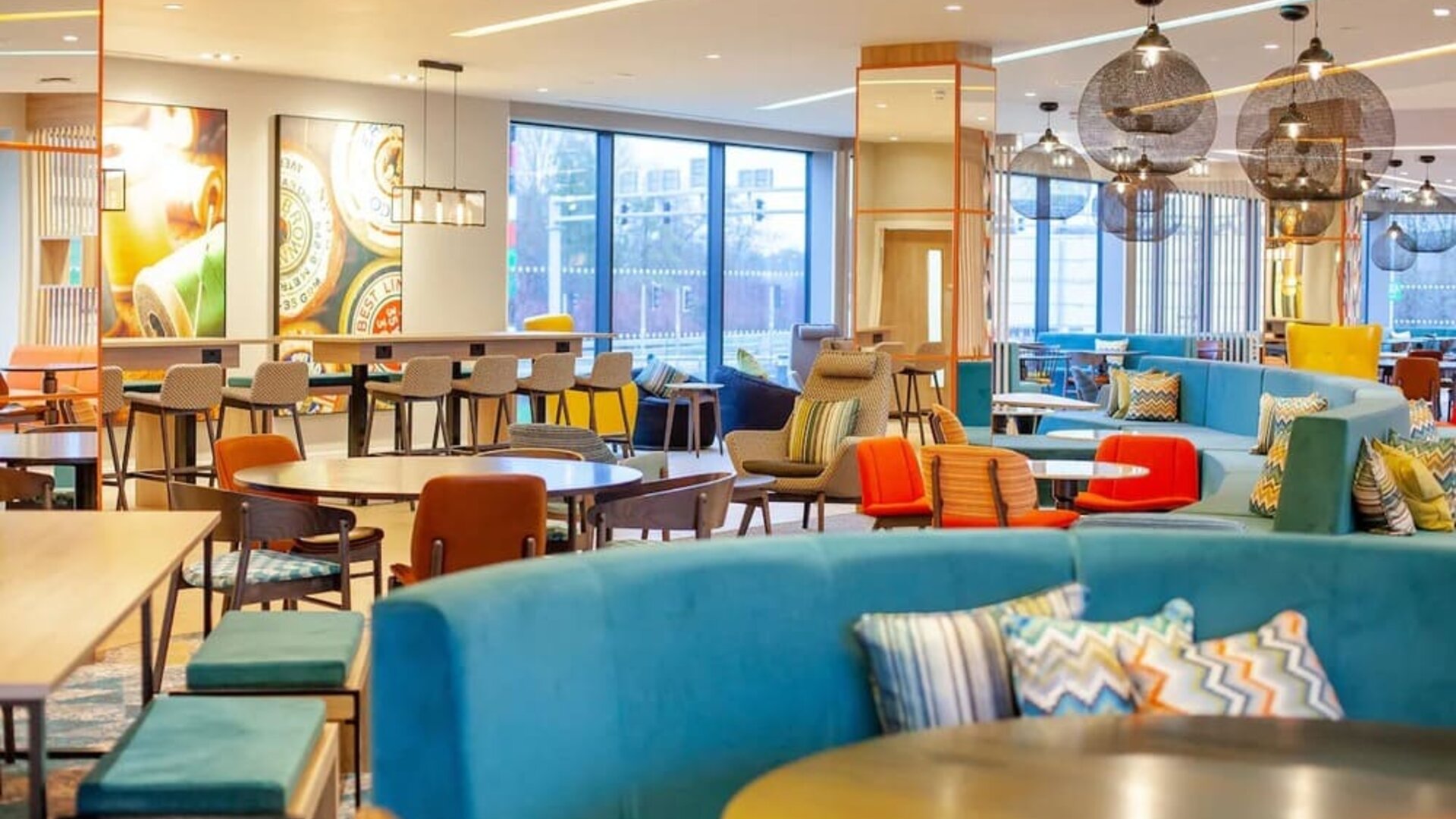 Colorful open-plan Hampton by Hilton hotel lounge with turquoise sofas, patterned cushions, assorted tables and chairs, contemporary pendant lights, large windows, and communal seating areas.