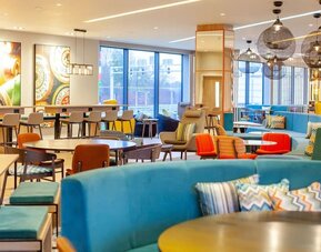 Colorful open-plan Hampton by Hilton hotel lounge with turquoise sofas, patterned cushions, assorted tables and chairs, contemporary pendant lights, large windows, and communal seating areas.