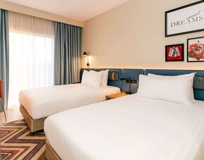 Bright contemporary hotel room with two beds, crisp white linens, blue upholstered headboard, framed wall art, floor-to-ceiling window with blue curtains, orange chair, wood flooring, and patterned rug.