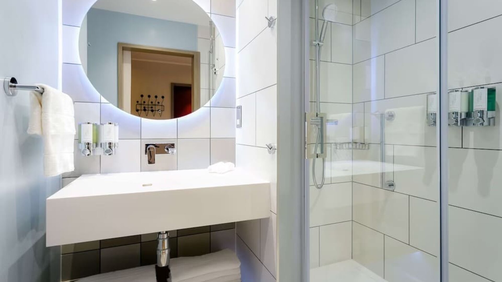 Bright contemporary hotel bathroom with a large illuminated round mirror, white rectangular sink, folded towels, glass-enclosed walk-in shower, handheld showerhead, and wall-mounted toiletries.