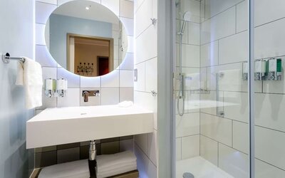 Bright contemporary hotel bathroom with a large illuminated round mirror, white rectangular sink, folded towels, glass-enclosed walk-in shower, handheld showerhead, and wall-mounted toiletries.