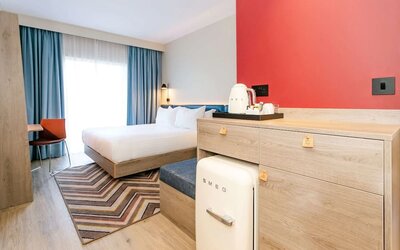 Modern hotel guest room with a king-size bed, white bedding, blue curtains, wood furnishings, red accent wall, compact SMEG refrigerator, tea and coffee station, patterned rug, and desk chair.