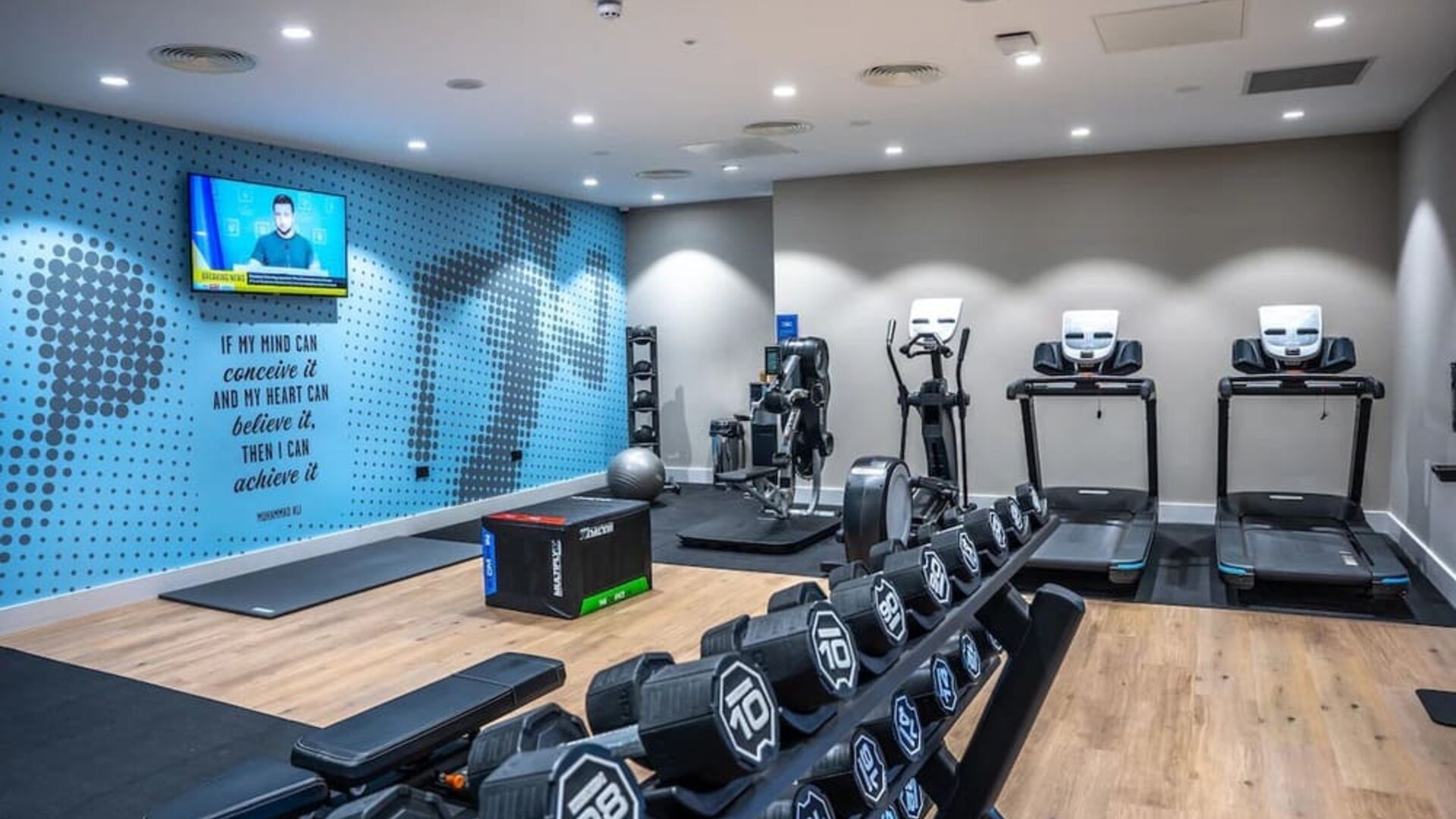 Modern hotel fitness center equipped with treadmills, an elliptical machine, exercise equipment, a dumbbell rack, workout bench, exercise ball, and wall-mounted television.