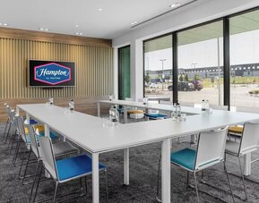 Bright Hampton by Hilton meeting room with white tables arranged in a U-shape, colorful chairs, bottled water, large windows with blinds, and a wall-mounted screen displaying the Hampton by Hilton logo.