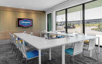 Bright Hampton by Hilton meeting room with white tables arranged in a U-shape, colorful chairs, bottled water, large windows with blinds, and a wall-mounted screen displaying the Hampton by Hilton logo.
