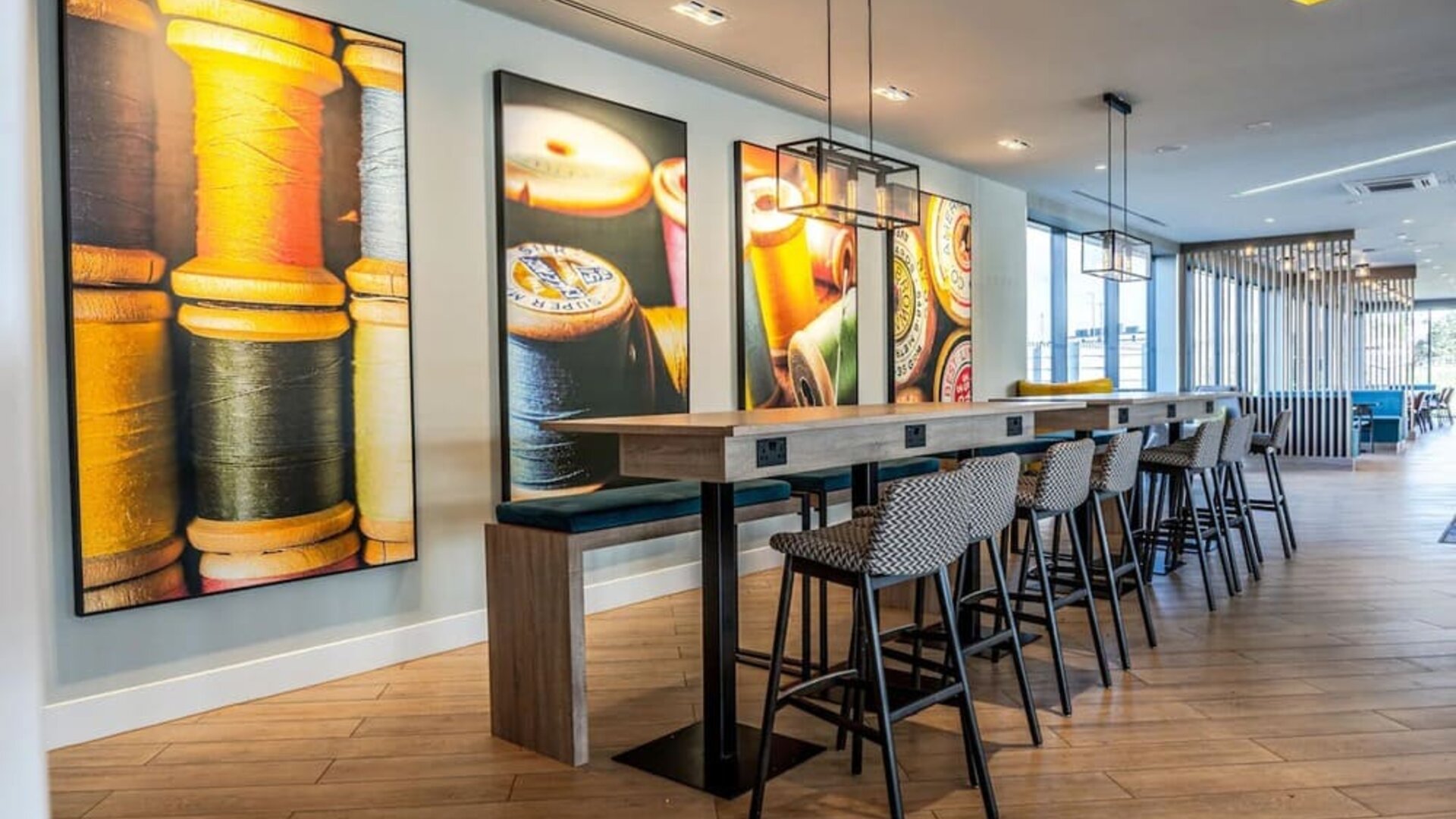 Contemporary hotel coworking space with a long communal wooden table, power outlets, patterned high stools, modern pendant lighting, and colorful wall artwork featuring spools of thread and textile imagery.