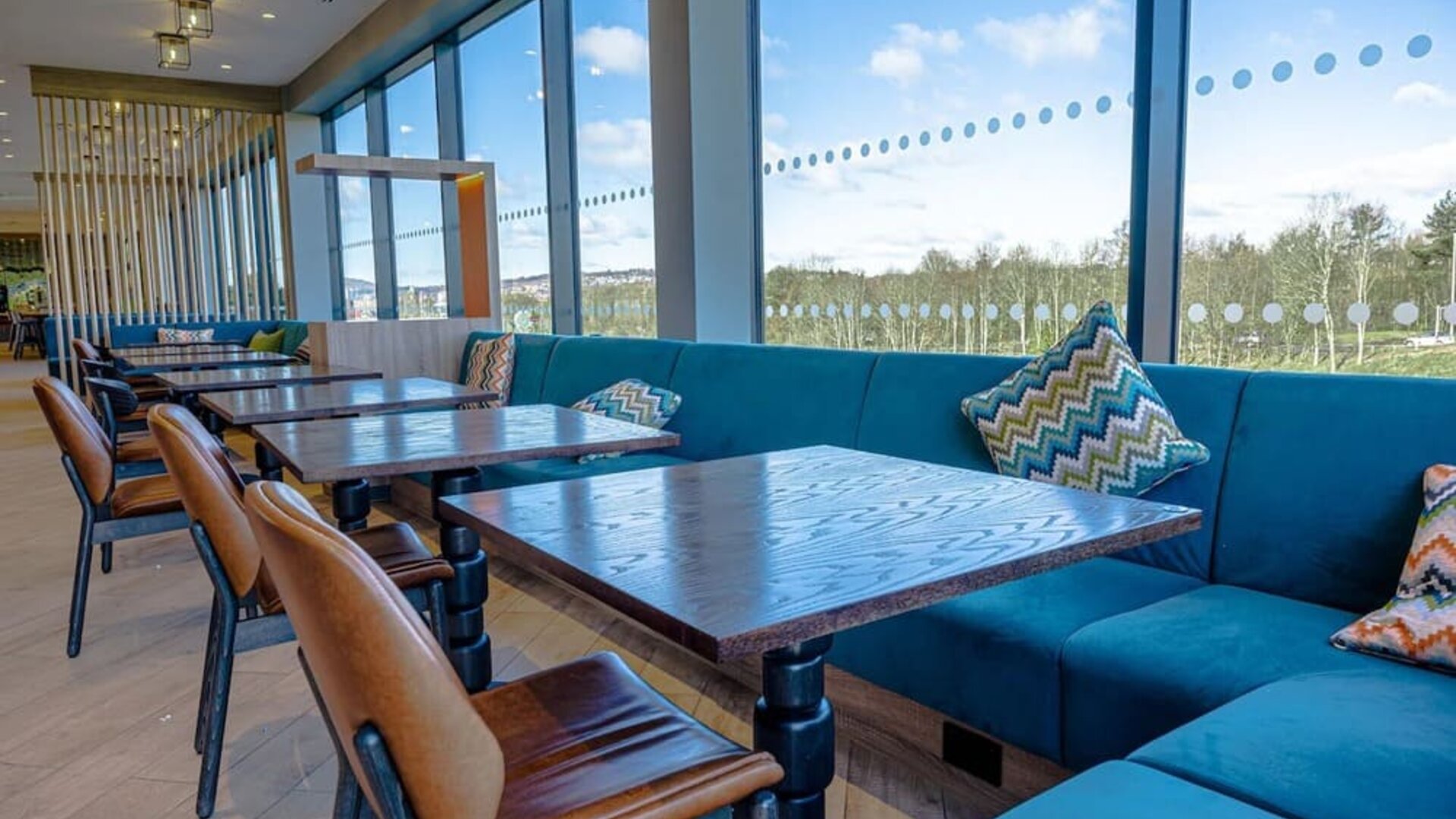 Spacious hotel dining area with large floor-to-ceiling windows, turquoise upholstered banquette seating, patterned cushions, dark wood tables, and brown leather dining chairs overlooking the surrounding landscape.