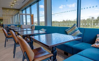 Spacious hotel dining area with large floor-to-ceiling windows, turquoise upholstered banquette seating, patterned cushions, dark wood tables, and brown leather dining chairs overlooking the surrounding landscape.