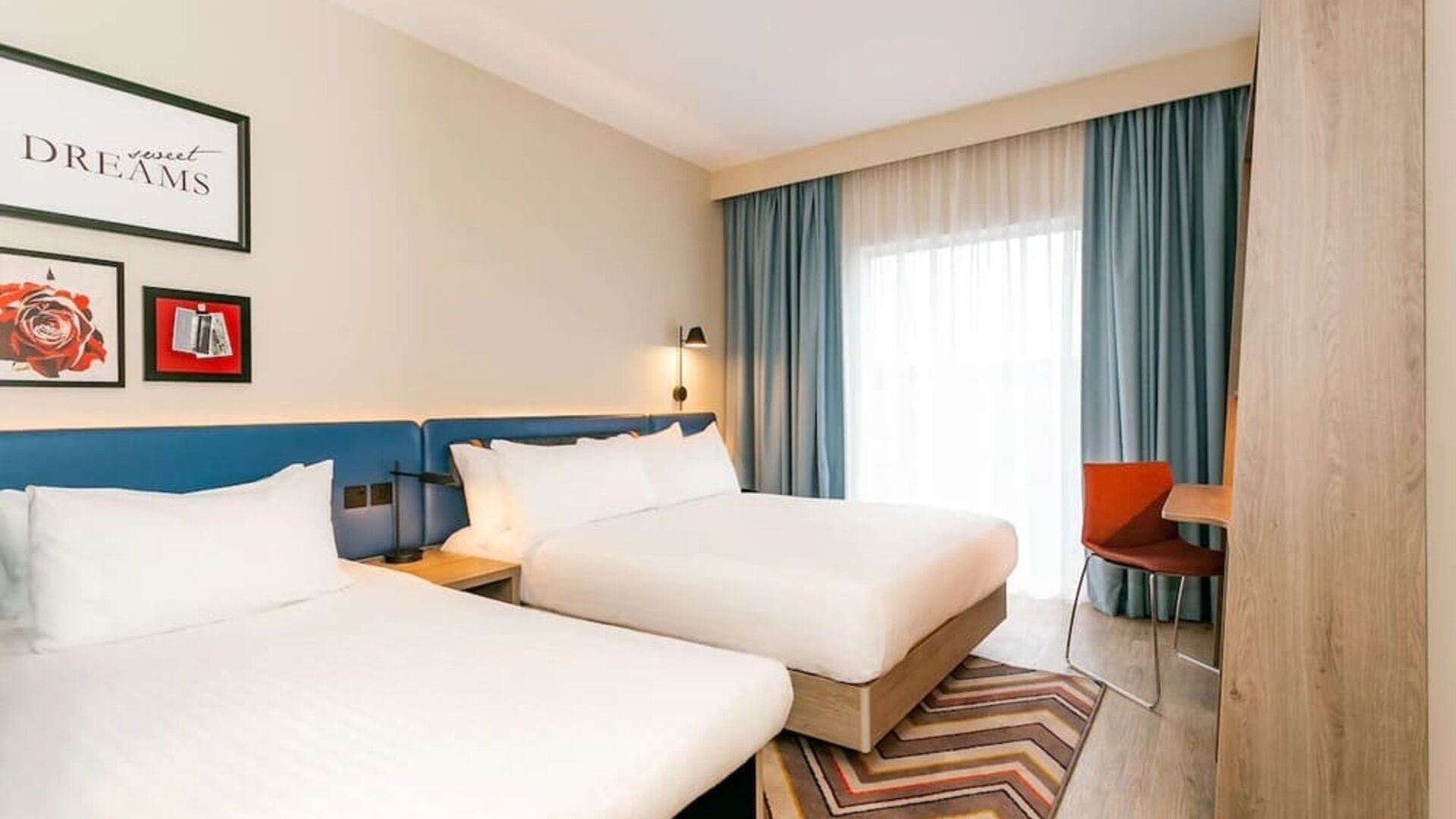 Bright modern hotel guest room with two beds dressed in white linens, a blue upholstered headboard, bedside lighting, floor-to-ceiling windows with blue curtains, a patterned rug, and a compact work desk.