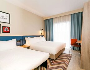 Bright modern hotel guest room with two beds dressed in white linens, a blue upholstered headboard, bedside lighting, floor-to-ceiling windows with blue curtains, a patterned rug, and a compact work desk.