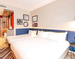 Contemporary hotel room with two beds dressed in white linens, long blue upholstered headboard, bedside lighting, framed artwork, patterned rug, tea and coffee station, and open wardrobe near the entrance.