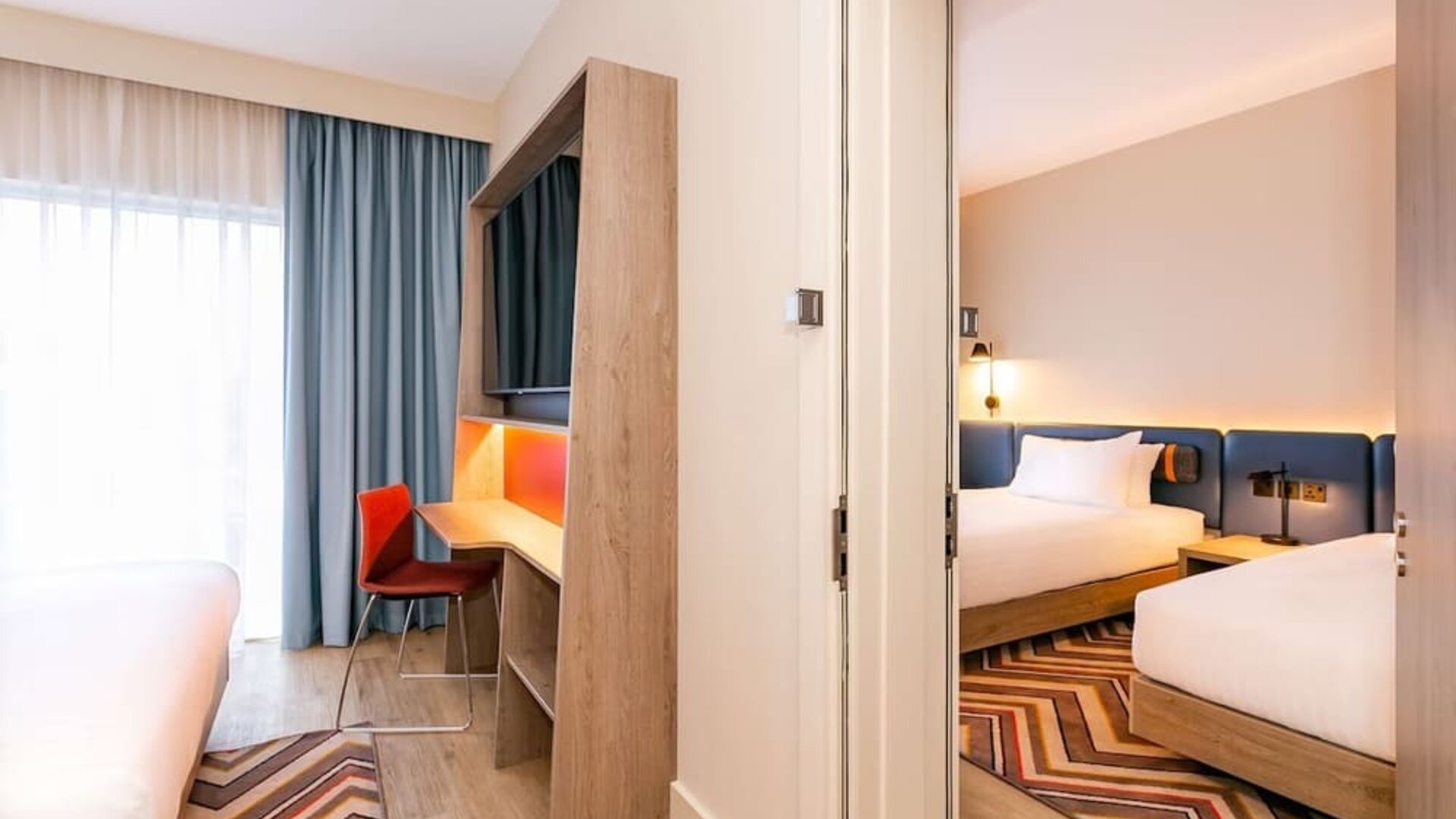 View between adjoining hotel sleeping areas featuring multiple beds with white linens, blue upholstered headboards, floor-to-ceiling windows with blue curtains, built-in desk and television unit, orange chair, wood flooring, and geometric rugs.