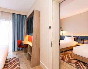 View between adjoining hotel sleeping areas featuring multiple beds with white linens, blue upholstered headboards, floor-to-ceiling windows with blue curtains, built-in desk and television unit, orange chair, wood flooring, and geometric rugs.