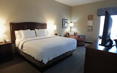 Spacious king guest room featuring a large bed with white linens, dark wood headboard, bedside lighting, upholstered chair, wall artwork, window and additional seating near the far wall.