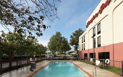 Hampton Inn outdoor swimming pool beside the hotel building, surrounded by a fenced concrete deck with lounge chairs, trees and parked vehicles under a bright daytime sky.