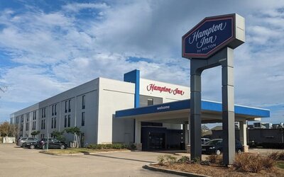 Daytime exterior of Hampton Inn by Hilton featuring the three-story hotel building, covered entrance, prominent roadside Hampton Inn sign, parked vehicles and a partly cloudy blue sky.