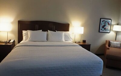 Hotel guest room with a king-size bed dressed in crisp white bedding, dark wood headboard, matching nightstands and lamps, plus an upholstered armchair and framed artwork.