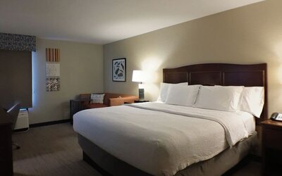 Spacious hotel guest room with a king-size bed dressed in white linens, dark wood headboard, bedside lamps, armchair, side table, wall artwork and television.