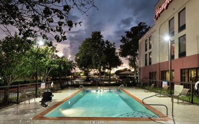 Illuminated outdoor hotel swimming pool at dusk with lounge chairs around the fenced pool deck, mature trees, parking area and the Hampton Inn building beneath a dramatic cloudy evening sky.