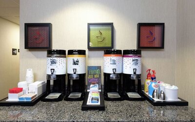 Hotel coffee station with four self-service beverage dispensers offering regular coffee, decaf and hot water, with stacked cups, tea selections, sweeteners and coffee creamers on a granite counter.