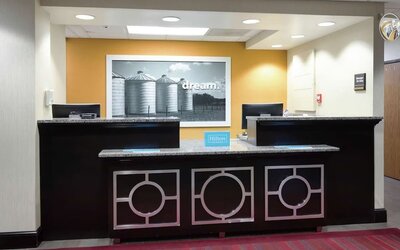 Hampton Inn reception area with two dark wood and granite front desk counters, modern geometric detailing and a large black-and-white wall photograph against a warm yellow accent wall.