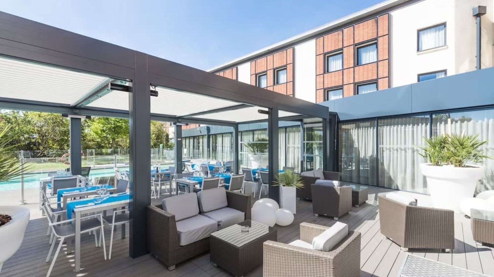 Outdoor terrace with lounge seating, dining tables, glass walls, and views of the hotel pool at Holiday Inn Toulouse Airport.
