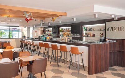Hotel bar with a long counter, bar stools, lounge seating, airplane decorations, and illuminated shelving at Holiday Inn Toulouse Airport.
