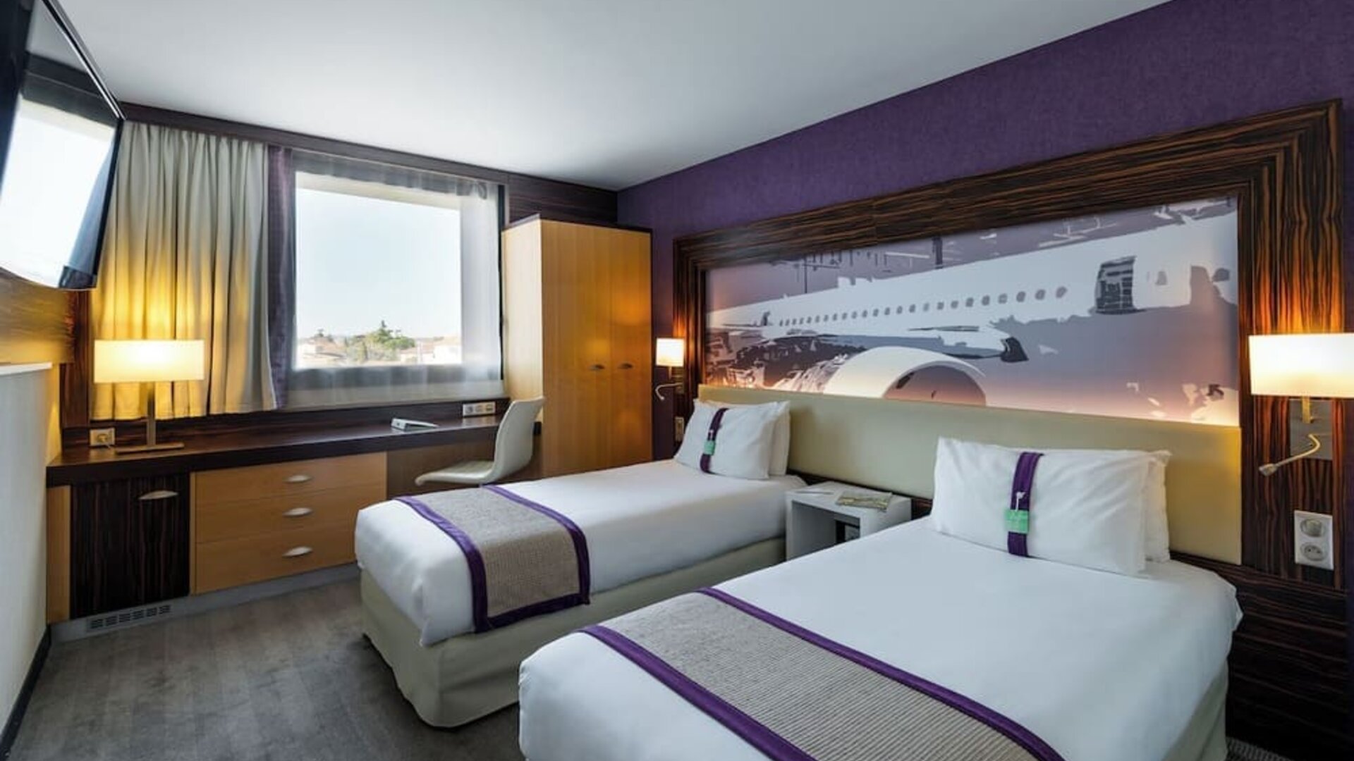 Guest room with two single beds, purple accents, airplane artwork, TV, work desk, and wardrobe at Holiday Inn Toulouse Airport.