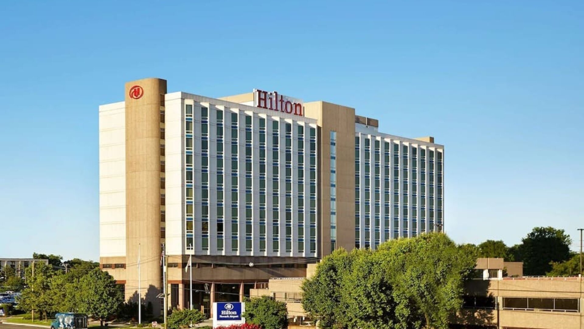 Exterior of Hilton Newark Airport with a multi-story facade, landscaped grounds, entrance driveway, and Hilton signage.