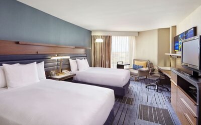 1. Guest room with two double beds, blue accent wall, bedside table, work desk, lounge chair, television, and large windows at Hilton Newark Airport.