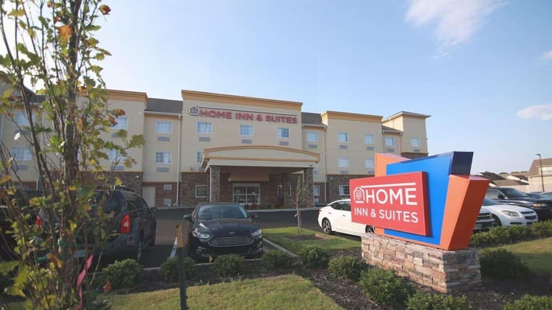 Exterior of At Home Inn & Suites showing the three-story hotel building, entrance, landscaped grounds, parking area, and hotel sign.