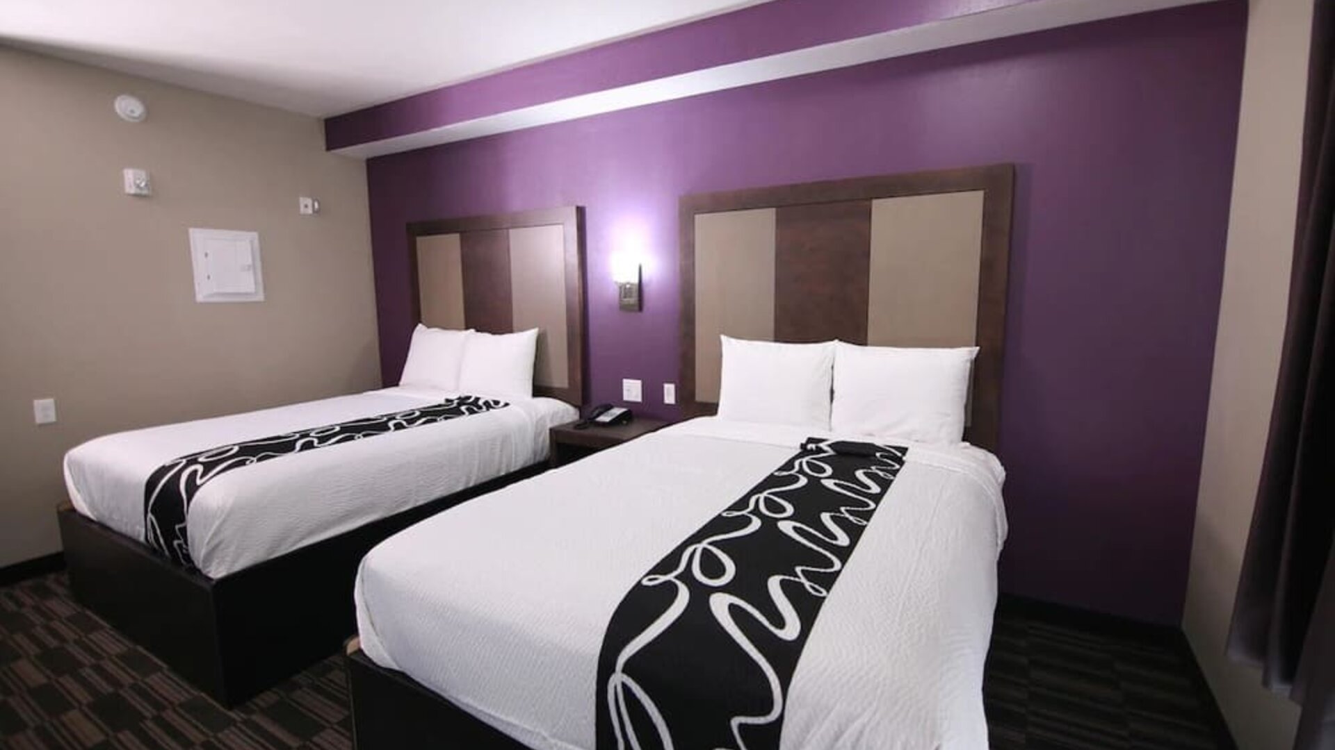 Guest room with two beds featuring white linens, dark headboards, patterned black bed runners, and a purple accent wall at At Home Inn & Suites.
