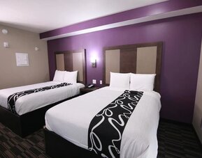Guest room with two beds featuring white linens, dark headboards, patterned black bed runners, and a purple accent wall at At Home Inn & Suites.