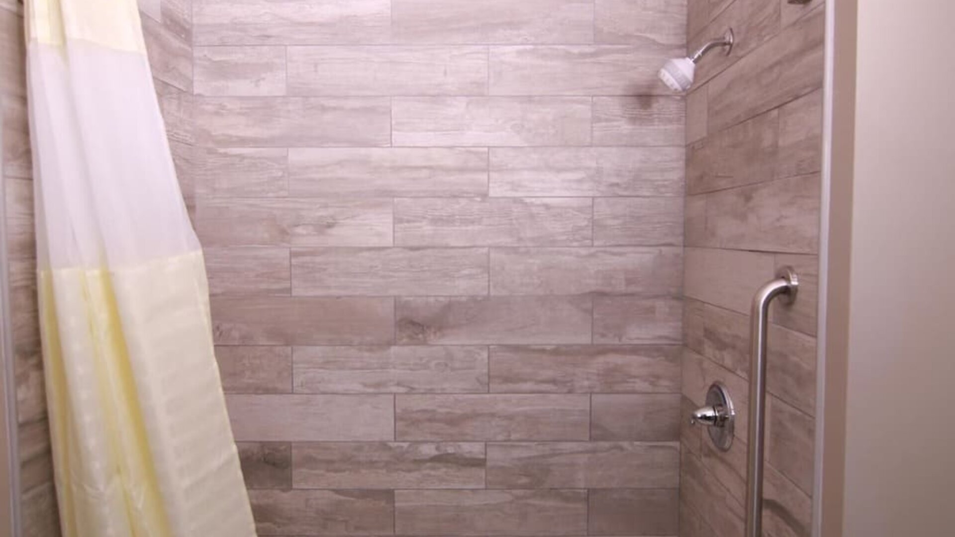 Bathtub and shower with wood-look tiled walls, curved shower rod, showerhead, grab bar, and white shower curtain at At Home Inn & Suites.