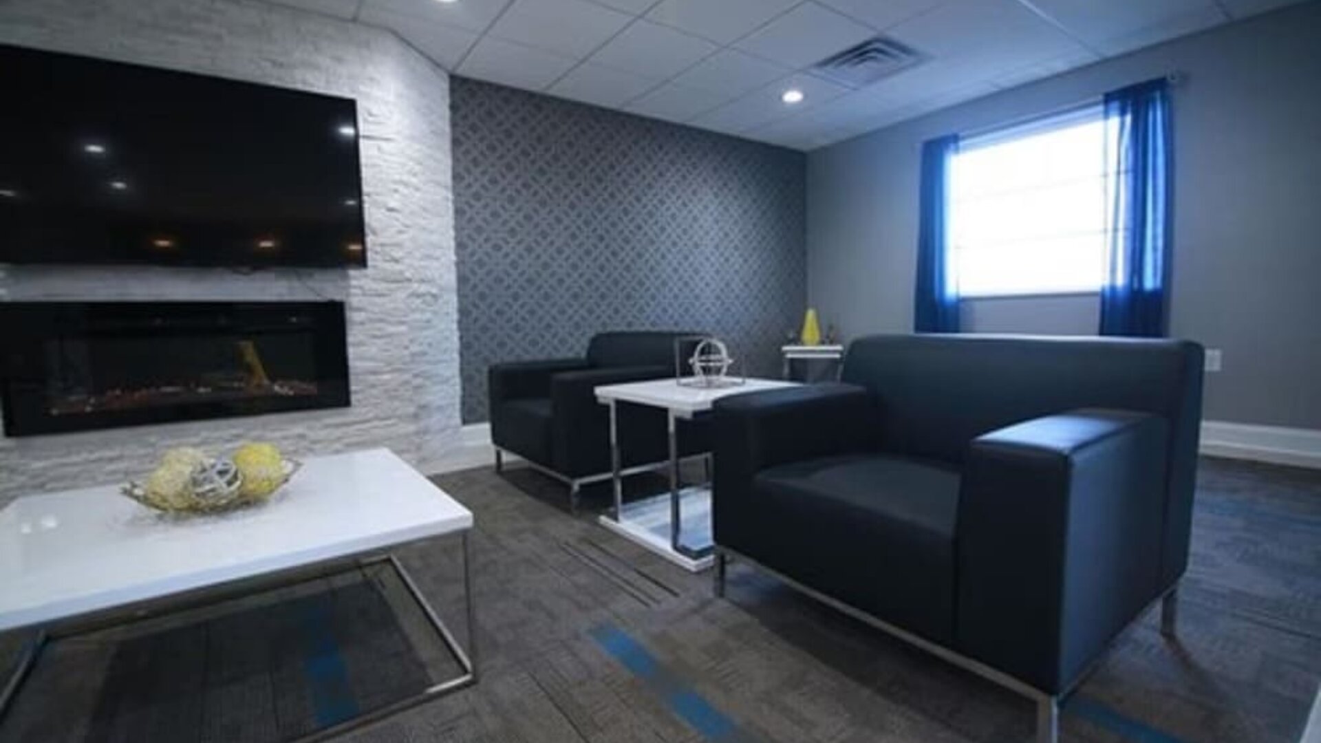 Hotel lounge seating area with black sofas, coffee tables, a fireplace, television, and large window at At Home Inn & Suites.