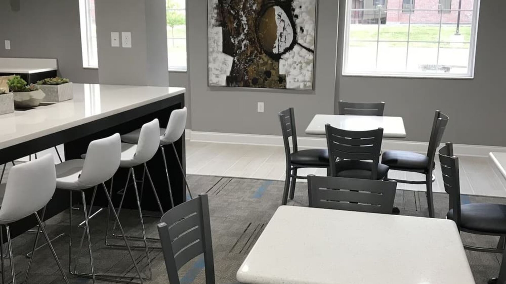 Hotel dining area with white tables, black chairs, bar-height seating, large windows, and contemporary artwork at At Home Inn & Suites.