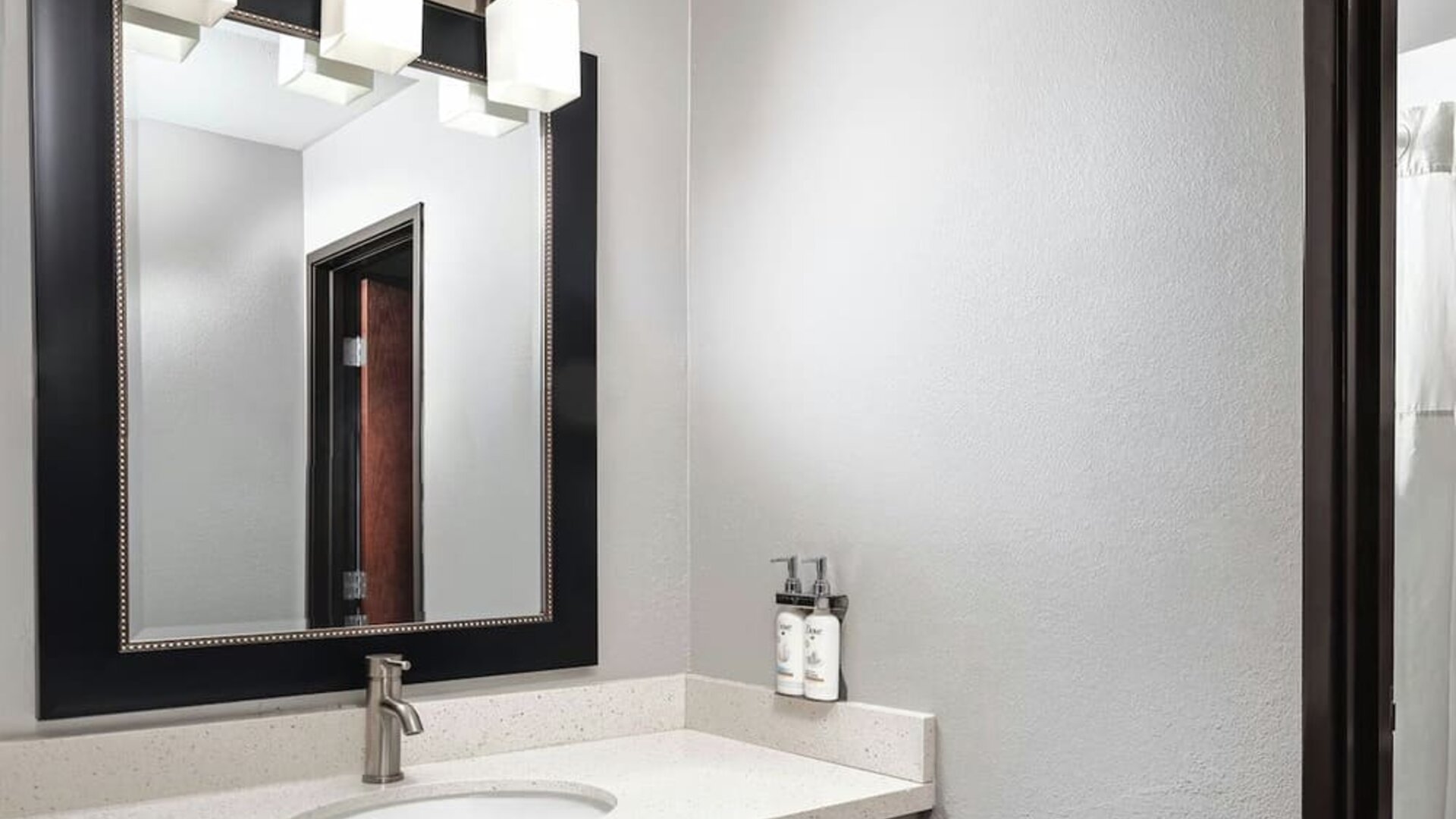 Bathroom vanity with a white countertop, oval sink, modern faucet, large framed mirror, and wall-mounted toiletries at Garner by IHG.