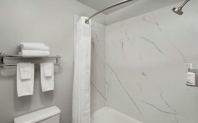 Bathroom with a bathtub and shower, white marble-look wall panels, curved shower rod, towels, and wall-mounted toiletries at Garner by IHG.