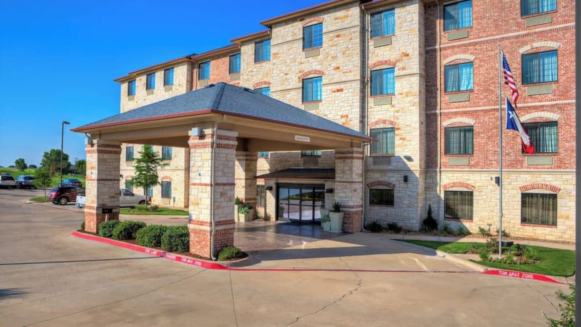 Front exterior of Holiday Inn Express & Suites Granbury showing the covered porte-cochère, landscaped grounds, parking area, and American and Texas flags.