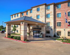 Front exterior of Holiday Inn Express & Suites Granbury showing the covered porte-cochère, landscaped grounds, parking area, and American and Texas flags.