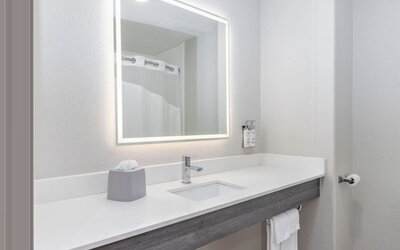 Bathroom vanity with a large illuminated mirror, white countertop, integrated sink, and wall-mounted toiletries at Holiday Inn Express & Suites Granbury.