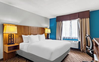 Bright hotel room with a king-size bed, white bedding, wooden headboard, bedside lamps, large window with curtains, work desk, and ergonomic chair.