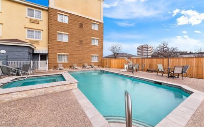 Hotel outdoor swimming pool and adjacent spa surrounded by a paved deck, lounge chairs, patio seating, wooden privacy fence, and multi-story hotel building.