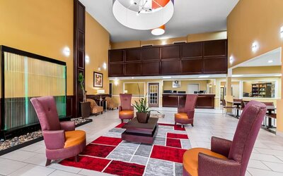 Spacious hotel lobby with colorful high-back lounge chairs, central coffee table, patterned area rug, indoor plants, reception desk, tiled flooring, and decorative water feature.
