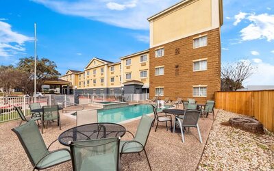 Outdoor hotel pool and patio area with tables, chairs and loungers surrounding the swimming pool and spa, with the hotel building and blue sky in the background.